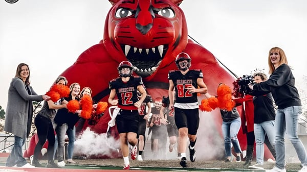 Athleten der Mena High School gehen durch Rauchwolken unter der großen roten Maskottchenfigur.