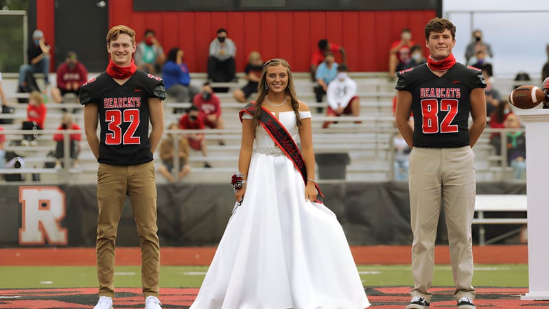 Eine Frau in weißem Kleid steht zwischen zwei Männern in Football-Trikots auf dem Sportfeld der Mena High School.