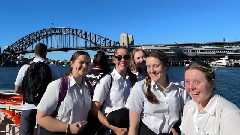 Eine Gruppe junger Schülerinnen der Menai High School posiert vor der Sydney Harbour Bridge mit der Stadtsilhouette im Hintergrund.