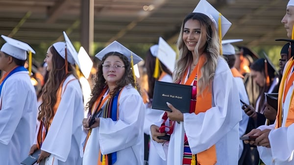 Eine Gruppe von Absolventinnen und Absolventen des Merced Union High School District steht in weißen Roben und Hüten mit Diplomen bei einer Zeremonie.