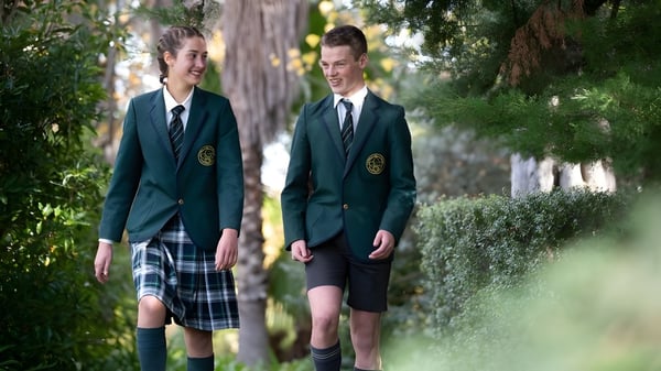 Zwei Schüler in Schuluniformen gehen zusammen auf einem Weg umgeben von üppigem Grün auf dem Campus des Mercedes College.