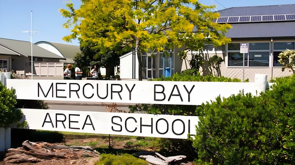 Das Schild der Mercury Bay Area School steht vor Bäumen mit einem Schulgebäude im Hintergrund.