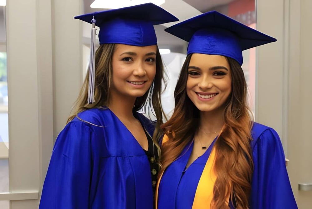 Zwei Absolventinnen in blauen Talaren und Hüten posieren lächelnd auf dem Campus des Mercy College.