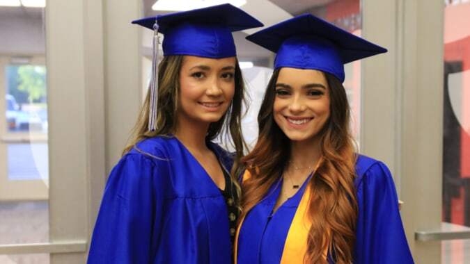 Zwei junge Frauen in blauen Abschlusshüten und Talaren stehen zusammen auf dem Mercy College Campus.
