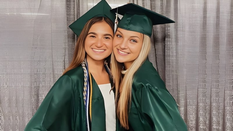 Zwei Schülerinnen der Mercyhurst Prep School in grünen Abschlussroben posieren vor einem grauen Vorhang.