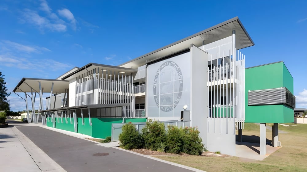 Das mehrstöckige Gebäude der Merrimac State High School mit weißen und grünen Fassaden und umliegender Landschaft unter blauem Himmel.