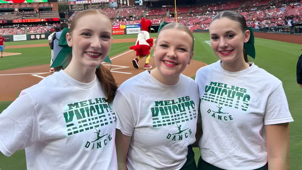 Drei Schülerinnen der Metro East Lutheran High School in weißen T-Shirts posieren lächelnd in einem Baseballstadion.
