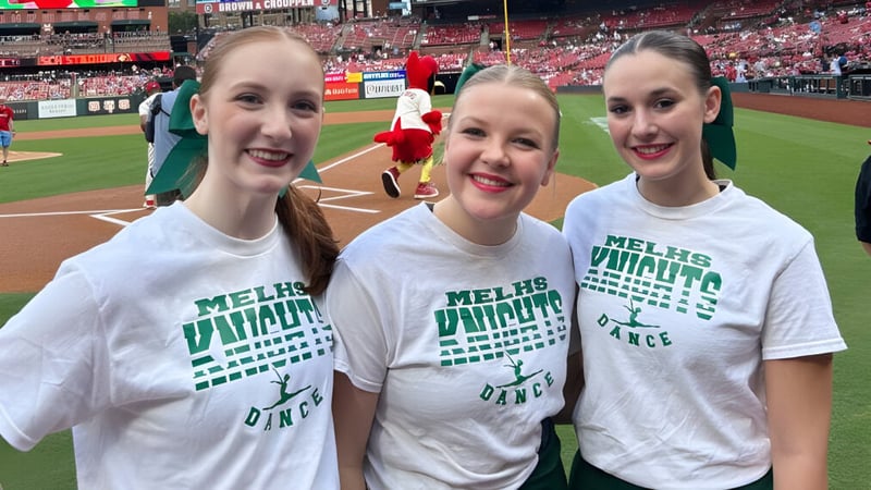 Drei Schülerinnen der Metro East Lutheran High School in weißen T-Shirts posieren lächelnd in einem Baseballstadion.