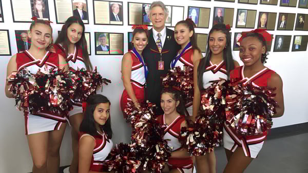 Eine Gruppe von Cheerleadern in roten und weißen Uniformen posiert vor einer Wand mit gerahmten Porträts an der Miami Beach Senior High School.