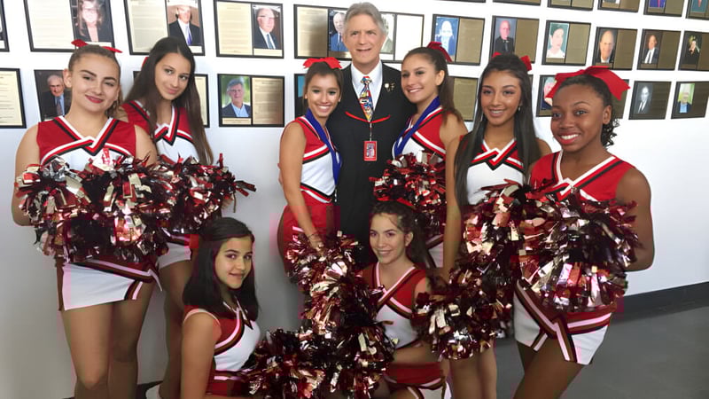Eine Gruppe von Cheerleadern in roten und weißen Uniformen posiert vor einer Wand mit gerahmten Porträts an der Miami Beach Senior High School.