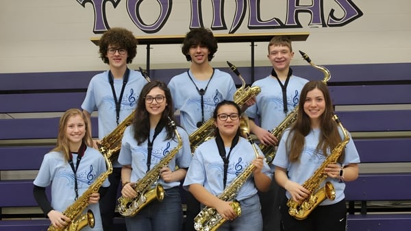 Eine Gruppe Schüler der Michigan Lutheran High School steht in einheitlichen Uniformen mit Saxophonen vor dem großen Tomcats-Schild.