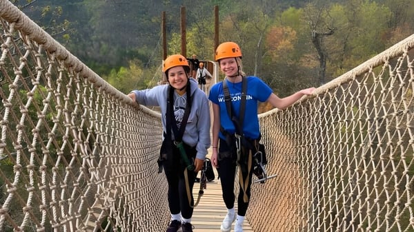 Zwei Personen mit Schutzhelmen überqueren eine Hängebrücke im Waldgebiet auf dem Gelände der Michigan Lutheran High School.