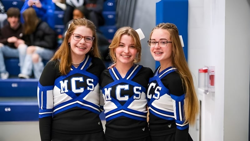 Drei Schülerinnen in Cheerleading-Uniformen mit MCS-Logo posieren vor blau-weißem Hintergrund auf dem Campus der Middletown Christian School.