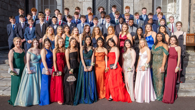 Eine große Gruppe von Schülerinnen und Schülern in formeller Kleidung steht vor einem Steinbau auf dem Campus des Midleton College.