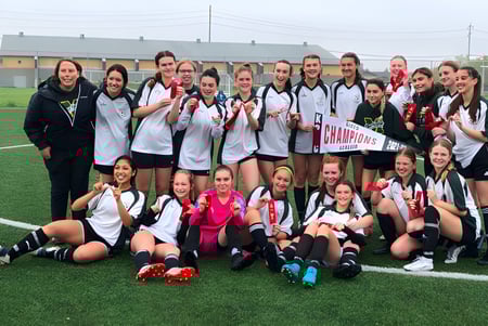 Eine Gruppe junger Fußballspielerinnen des Miles Macdonell Collegiate feiert auf dem Spielfeld mit einem Meisterschaftsbanner ihren Sieg.