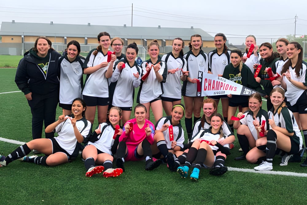 Eine Gruppe junger Fußballspielerinnen des Miles Macdonell Collegiate feiert auf dem Spielfeld mit einem Meisterschaftsbanner ihren Sieg.
