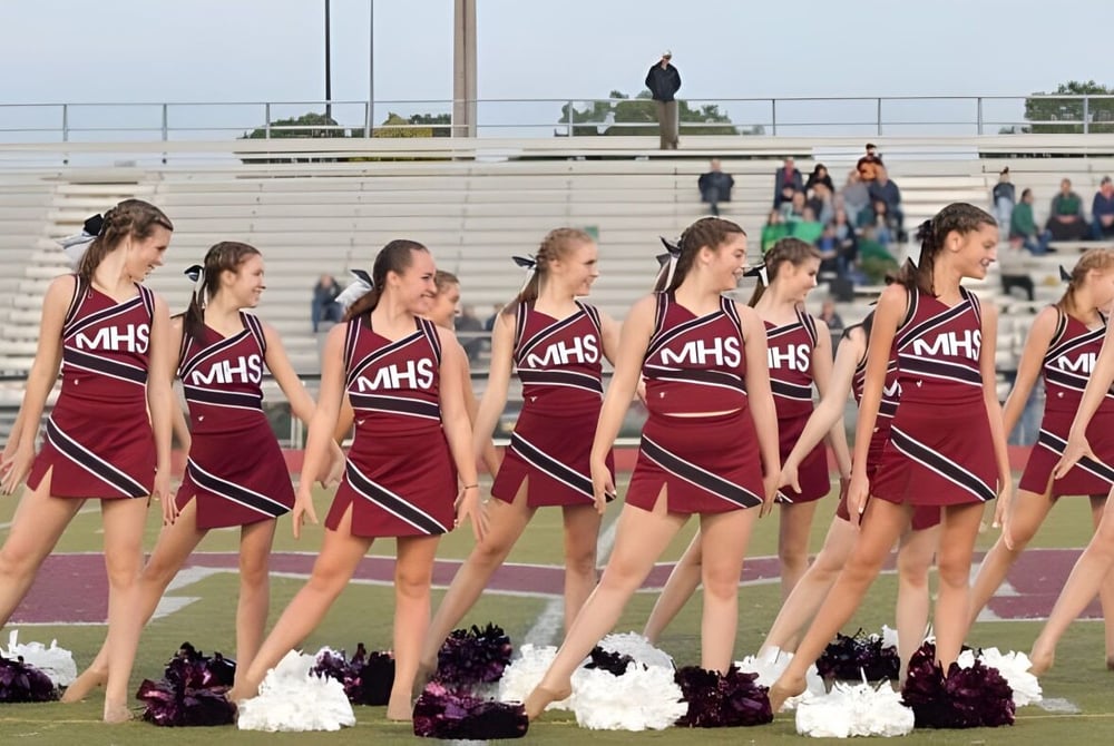Eine Gruppe Cheerleader in passenden Uniformen steht auf dem Sportfeld des Milford Public School Districts vor Zuschauern.