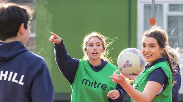Zwei Schülerinnen in Sportuniform mit Rugbyball sprechen auf dem Gelände von Mill Hill International mit einem Mann im Hintergrund.