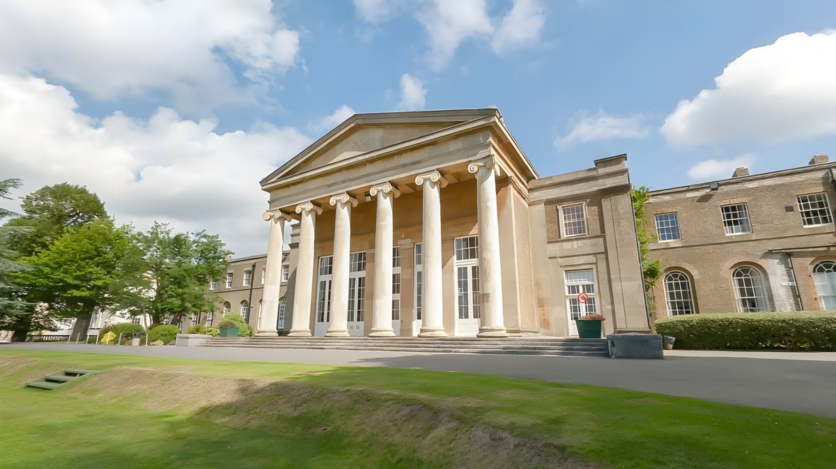 Das klassische Schulgebäude der Mill Hill School mit Säulen und einem Rasen im Vordergrund unter bewölktem Himmel.