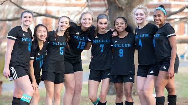 Eine Gruppe junger Athletinnen der Miller School of Albemarle posiert gemeinsam draußen vor Bäumen und Gebäuden.