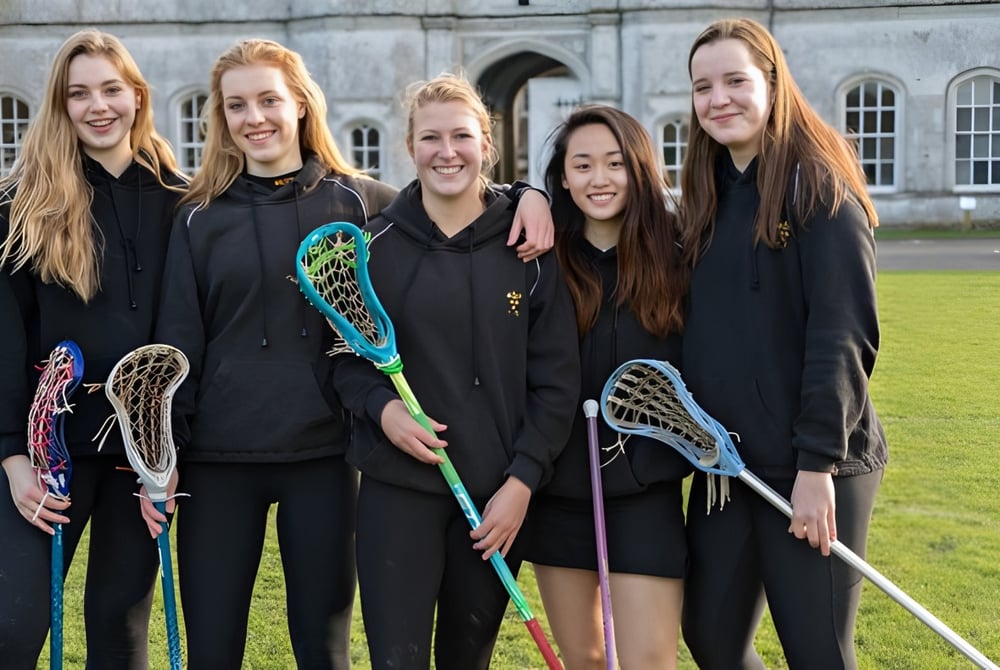 Eine Gruppe Schülerinnen der Milton Abbey School steht mit Lacrosse-Schlaegern vor einem historischen Gebaeude mit Rundbogenfenstern.
