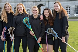 Eine Gruppe Schülerinnen der Milton Abbey School steht mit Lacrosse-Schlägern vor einem historischen Gebäude mit Rundbogenfenstern.