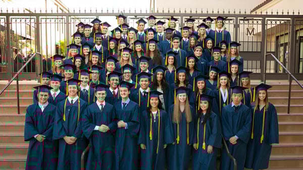 Eine große Gruppe von Absolventinnen und Absolventen steht in blauen und goldenen Talaren auf den Stufen eines Gebäudes auf dem Campus der Mission College Preparatory Catholic High School.