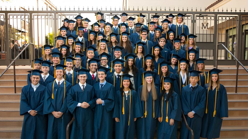 Eine große Gruppe von Absolventinnen und Absolventen steht in blauen und goldenen Talaren auf den Stufen eines Gebäudes auf dem Campus der Mission College Preparatory Catholic High School.