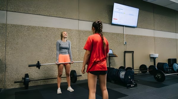 Zwei Frauen im Sportoutfit stehen in der Sporthalle der Mission Secondary School vor Trainingsgeräten und einem Bildschirm.
