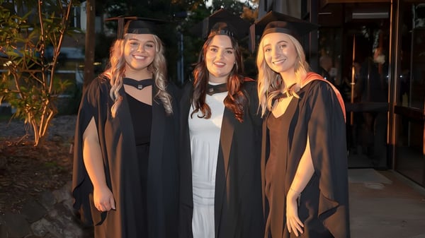 Drei Schülerinnen der Mitchelton State High School stehen in Abschlusstracht draußen vor Bäumen und Gebäuden.