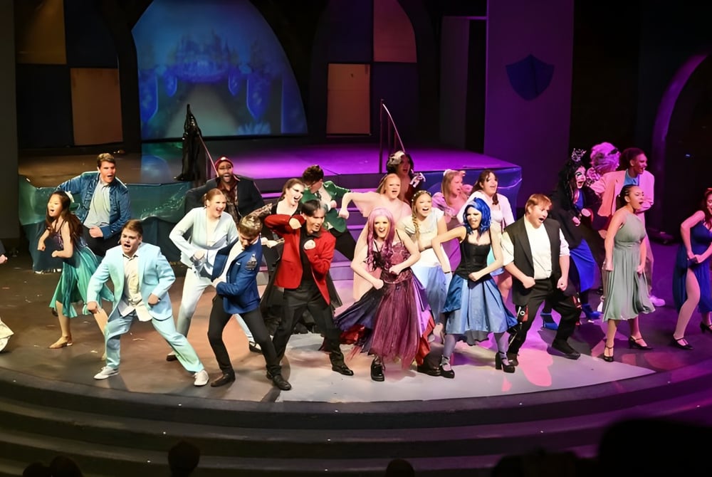 Eine Gruppe von Schülerinnen und Schülern in bunten Kostümen steht auf der Bühne der M.M. Robinson High School vor einem abstrakten Hintergrund.