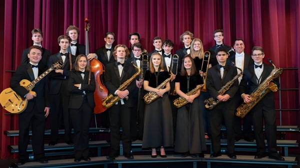 Eine Gruppe von Musikern des Moate Community College steht in formeller Kleidung mit Instrumenten auf einer Bühne vor rotem Vorhang.