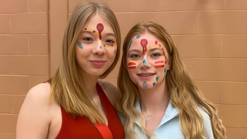 Zwei Schülerinnen der Mobile Central High mit farbiger Gesichtsbemalung lächeln vor einer beige Wand.