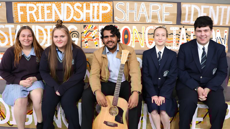 Eine Gruppe von Schülerinnen und Schülern steht vor einem bunten Graffiti mit den Worten Friendship, Share und Identity auf dem Campus der Modbury High School.