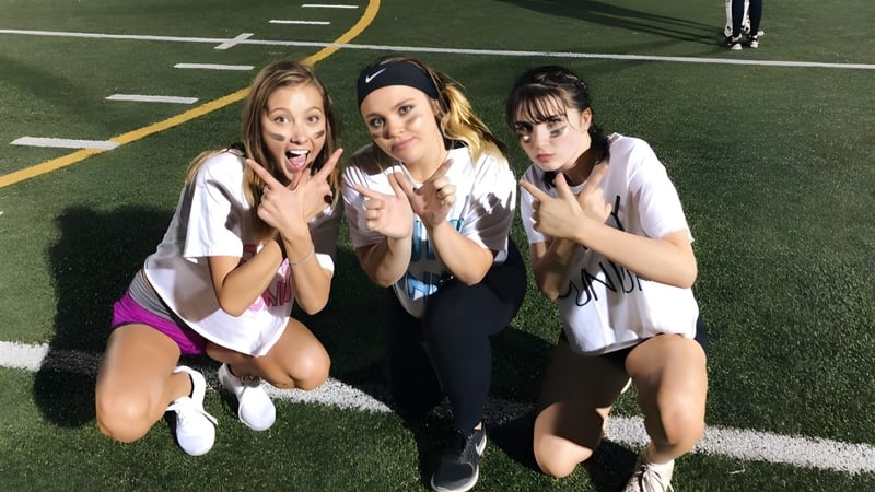 Drei Schülerinnen der Monarch High School posieren auf dem Sportplatz mit Spielfeldmarkierungen und Toren im Hintergrund.
