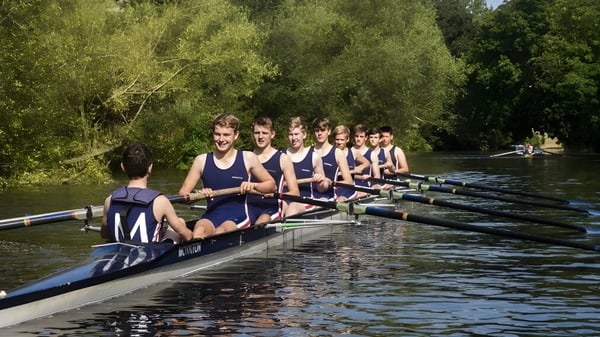Eine Gruppe Schüler der Monkton Combe School sitzt in einem Ruderboot auf einem ruhigen Fluss vor grüner Baumkulisse.