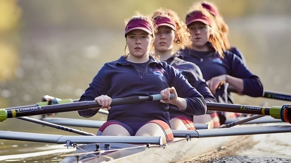 Eine Gruppe von Schülerinnen der Monmouth School for Girls rudert gemeinsam auf dem Wasser vor einer naturnahen Landschaft.