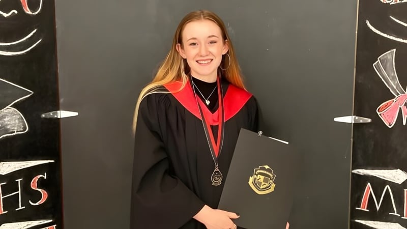 Eine junge Frau in Abschlussrobe steht vor einer Tafel mit Zeichnungen auf dem Campus der Montague Regional High School.