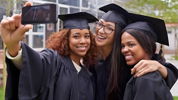 Drei Absolventinnen der Montcalm Secondary School posieren in ihren Abschlussgewändern für ein Selfie vor einem Schulgebäude mit Bäumen.