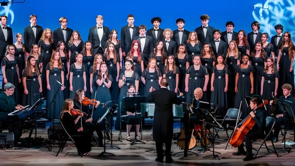 Ein großer Chor und ein Orchester führen unter Leitung eines Dirigenten auf der Bühne der Monte Vista Christian School vor blauem Hintergrund ein Musikstück auf.