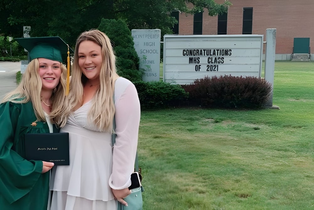 Zwei Schülerinnen in Abschlussroben stehen vor einem Schild mit Glückwünschen zur Klasse 2022 auf dem Campus der Montpelier High School.