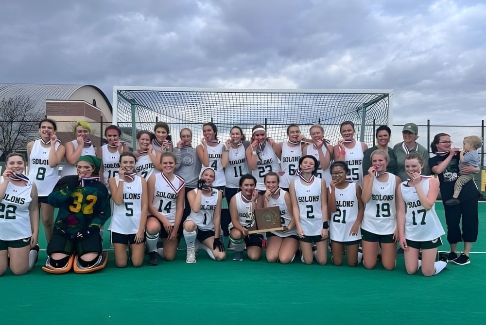 Eine Gruppe junger Sportlerinnen posiert auf dem Hockeyfeld des Montpelier Roxbury Public School District bei bewölktem Himmel.