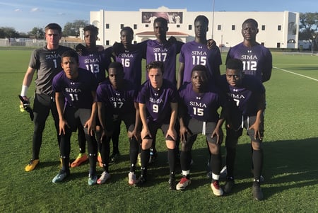 Eine Gruppe von Fußballspielern in lila Trikots steht auf dem Spielfeld der Montverde Academy mit einem Gebäude im Hintergrund.
