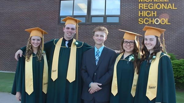 Eine Gruppe von Absolventinnen und Absolventen der Morell Regional High School in grünen Talaren steht vor dem Schulgebäude mit einem Schuladministrator.