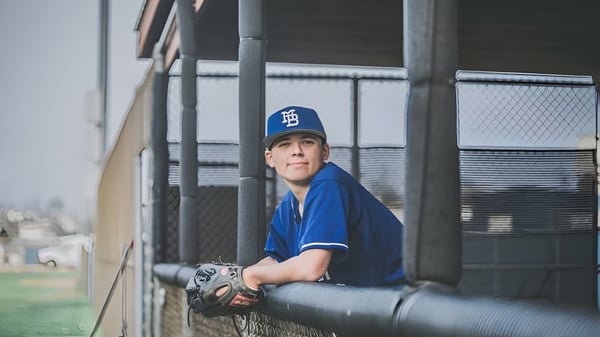 Ein junger Baseballspieler der Morro Bay High School steht in blauer Uniform hinter einem Maschendrahtzaun mit Baseballfeld im Hintergrund.