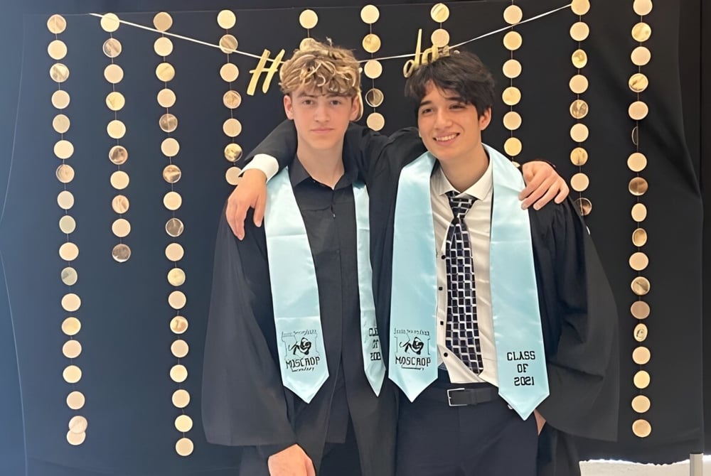 Zwei junge Männer in formeller Kleidung stehen vor einem dekorativen Hintergrund auf einer Veranstaltung der Moscrop School.