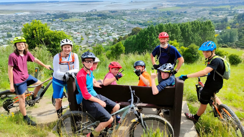 Eine Gruppe von Schülerinnen und Schülern der Motueka High School in bunter Fahrradausrüstung posiert auf einem Bergweg mit Blick auf die Stadt im Hintergrund.