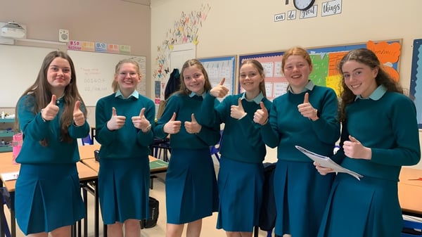 Sechs Schülerinnen in einheitlichen Uniformen stehen in einem farbenfrohen Klassenzimmer auf dem Campus der Mount Anville Secondary School und zeigen Daumen hoch.