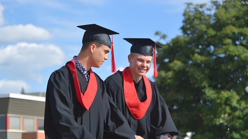 Zwei Schüler der Mount Baker Secondary School stehen in ihren Abschlussroben im Freien vor Bäumen und blauem Himmel.