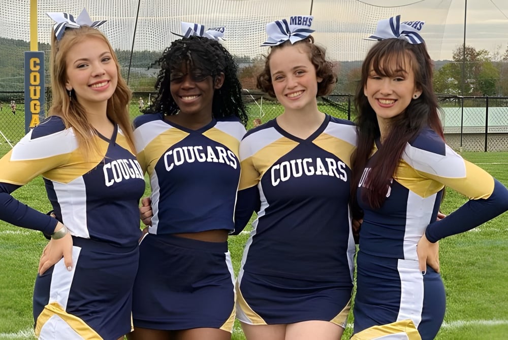 Vier Schülerinnen der Mount Blue High School posieren gemeinsam auf dem Sportfeld mit Bergen im Hintergrund.